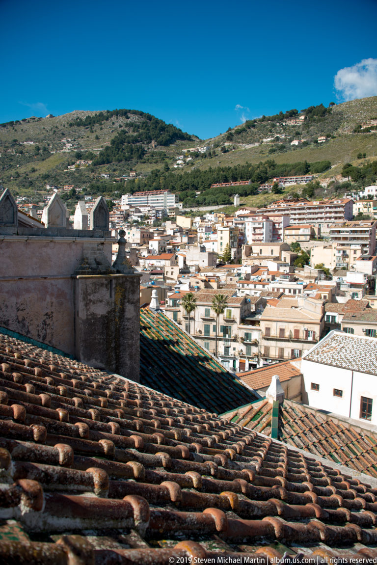 Dark tiled roofs overlook closely packed hillside houses and green slopes in Monreale.