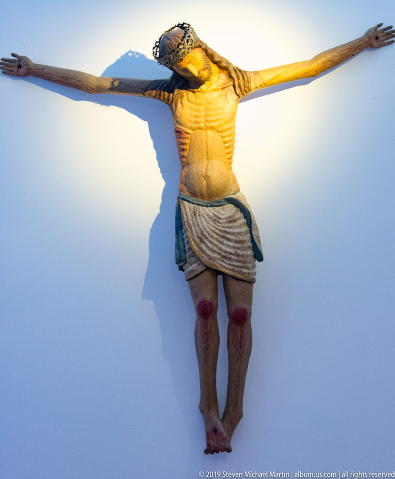 A carved figure of Christ with outstretched arms and a pale loincloth is viewed from below against the sky.