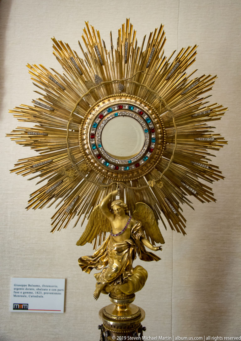 A gold sunburst reliquary with a central oval and small sculpted figure stands against a pale display wall.