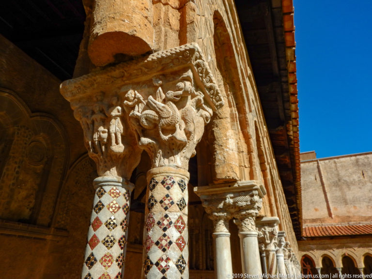 A carved capital and patterned columns frame the sunlit wall and roof beyond the cloister arcade.