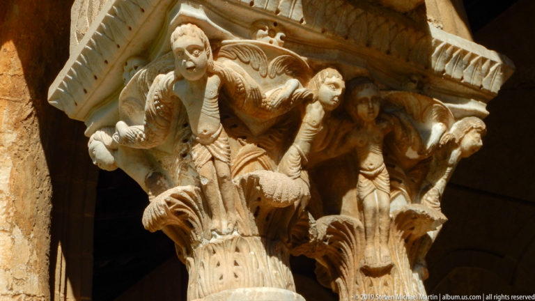A robed figure and smaller carved forms decorate the underside of a cloister capital.