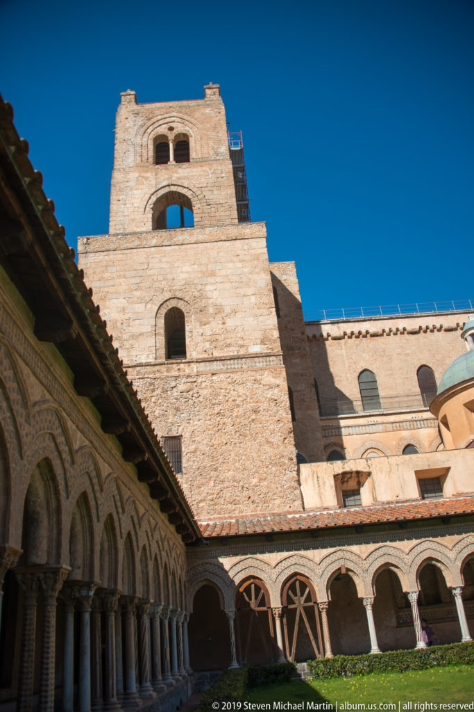A square stone bell tower rises beyond a shaded cloister arcade and garden.