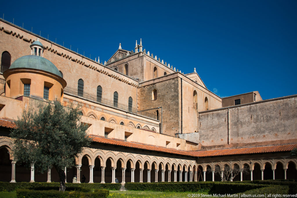 Pale cathedral buildings with roof pinnacles rise above the cloister’s long arcaded wall.