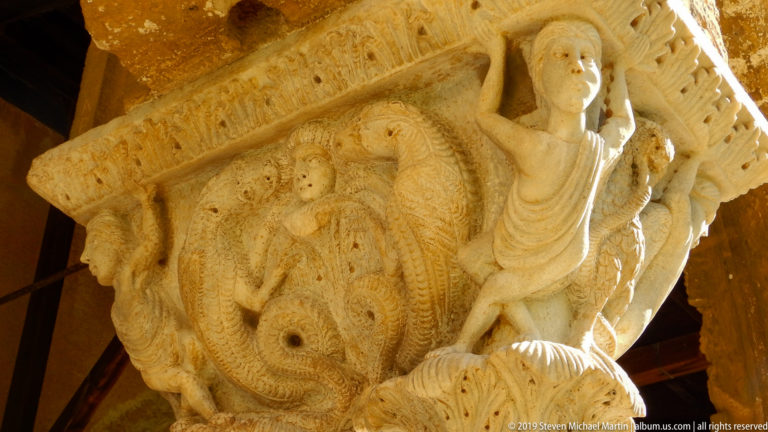 Stone capital with a close-packed group of carved figures beneath a projecting abacus.