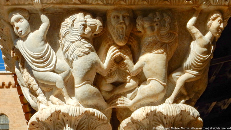 A multi-figure stone capital projects from the shadowed cloister arcade.