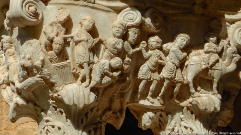 Carved figures in varied poses wrap around a stone capital beneath a projecting abacus.