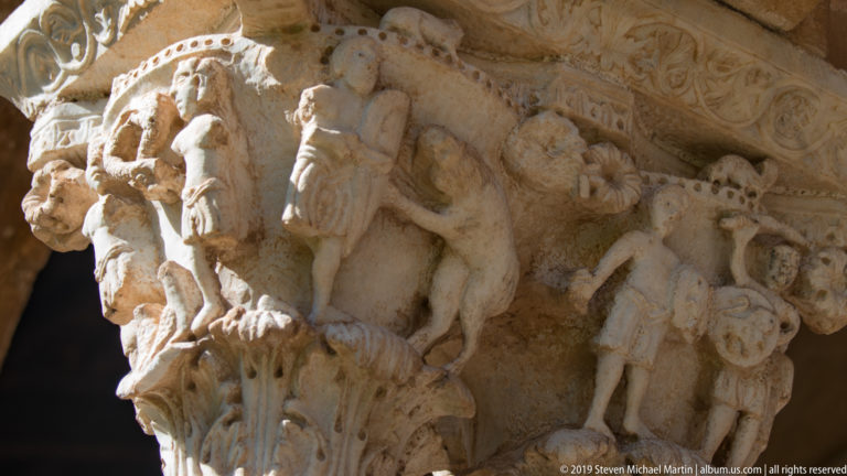 A row of carved figures surrounds an upright form on a stone cloister capital.