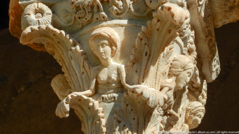 A central robed figure and surrounding relief are carved into a cloister capital.
