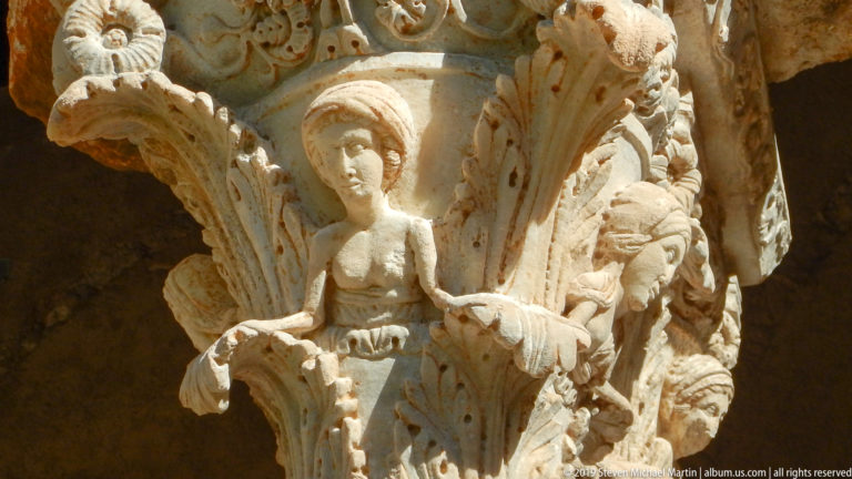 A standing robed figure is carved among smaller forms on a stone capital.