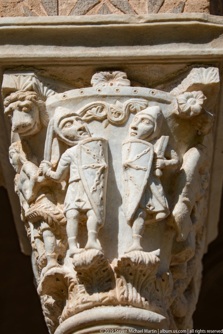A close, upward view of a stone capital with layered figures and foliage in deep shadow.