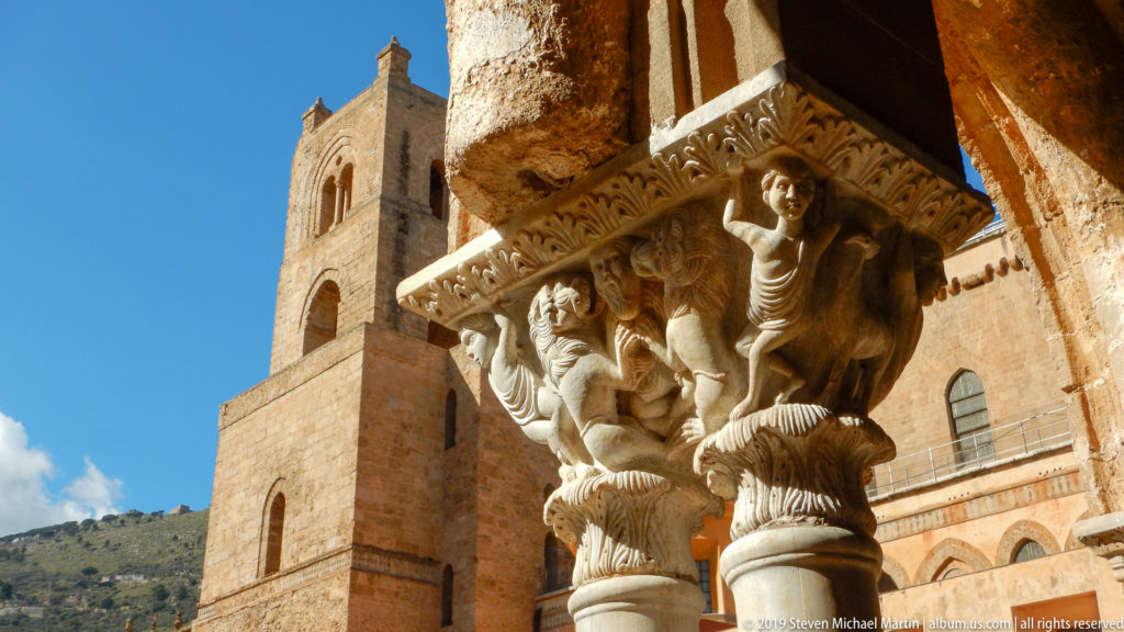 A carved cloister capital stands in the foreground with the cathedral tower behind it.