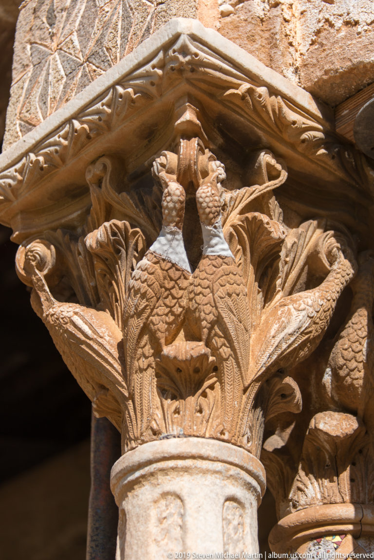 A sculpted capital with grouped figures beneath an overhanging stone abacus.
