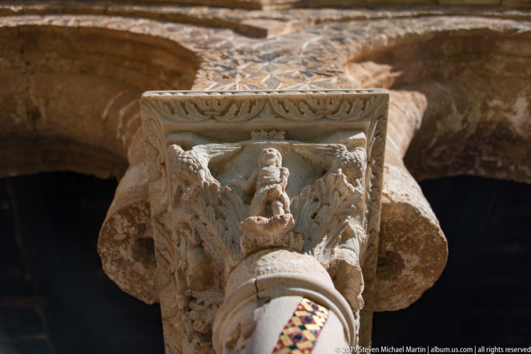 A small colored figure appears within a stone capital surrounded by carved figures and architecture.