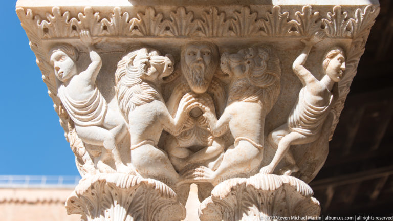 Two standing figures and smaller forms are carved into a cloister capital beneath a patterned band.