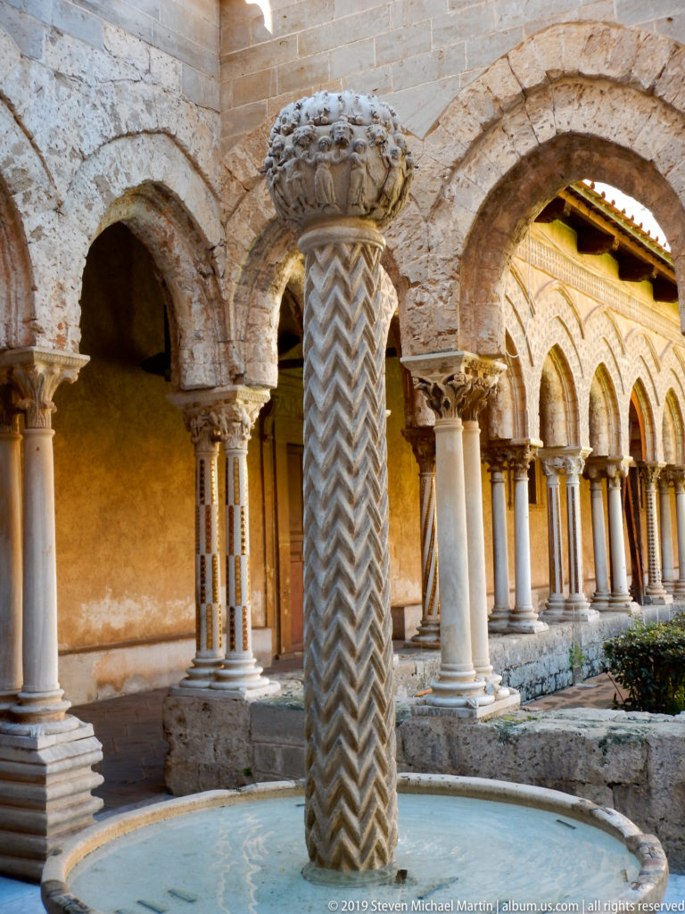 A carved fountain bowl and zigzag-patterned shaft rise from a circular basin in the cloister.