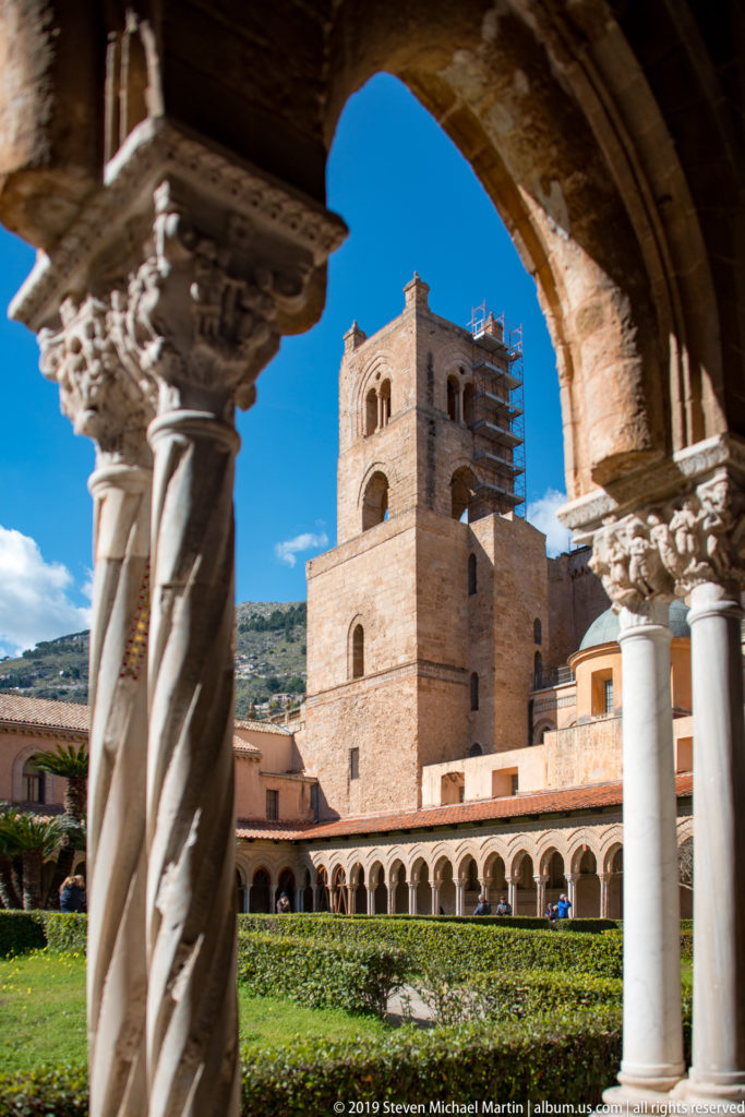 A square tower rises beyond two foreground columns and a shaded cloister arch.
