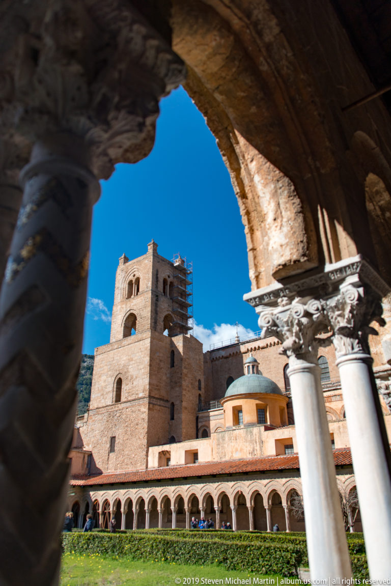 A stone tower and adjoining roofs seen through a cloister arch, with the arcade below.
