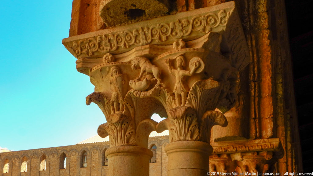 A stone cloister capital with small human figures and foliage carved beneath a band of roundels.
