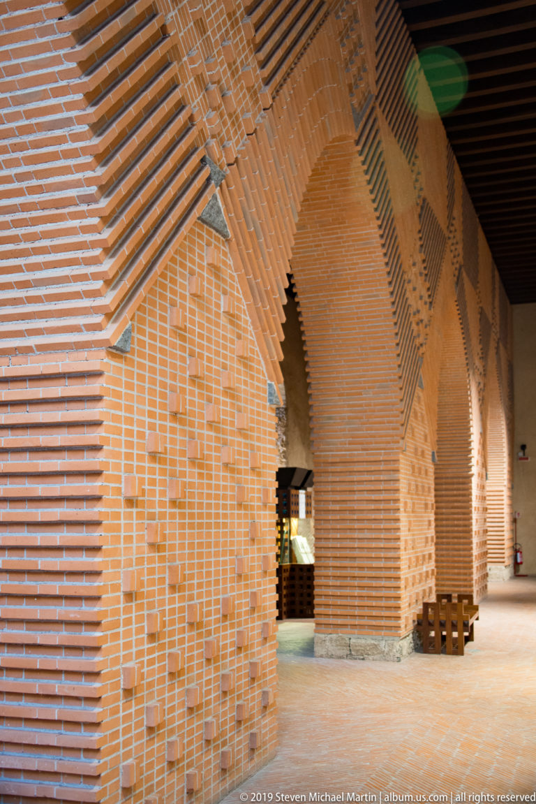 A shadowed brick passage with a tiled roof opens onto a sunlit area and low wooden barrier.