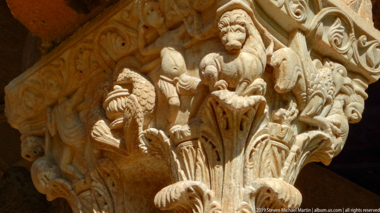 A raised-arm figure stands among smaller forms on a carved cloister capital.