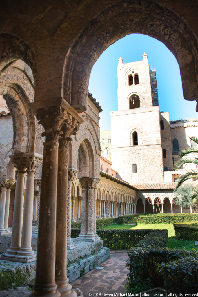 A square stone tower rises above Monreale’s cloister arcades and garden, seen through a shaded arch.