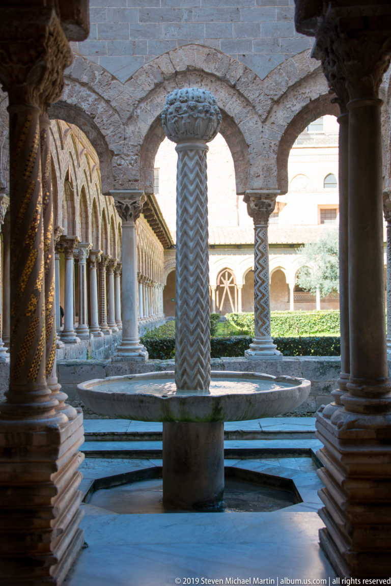 A cloister fountain with a patterned central shaft, framed by shadowed arches and columns.