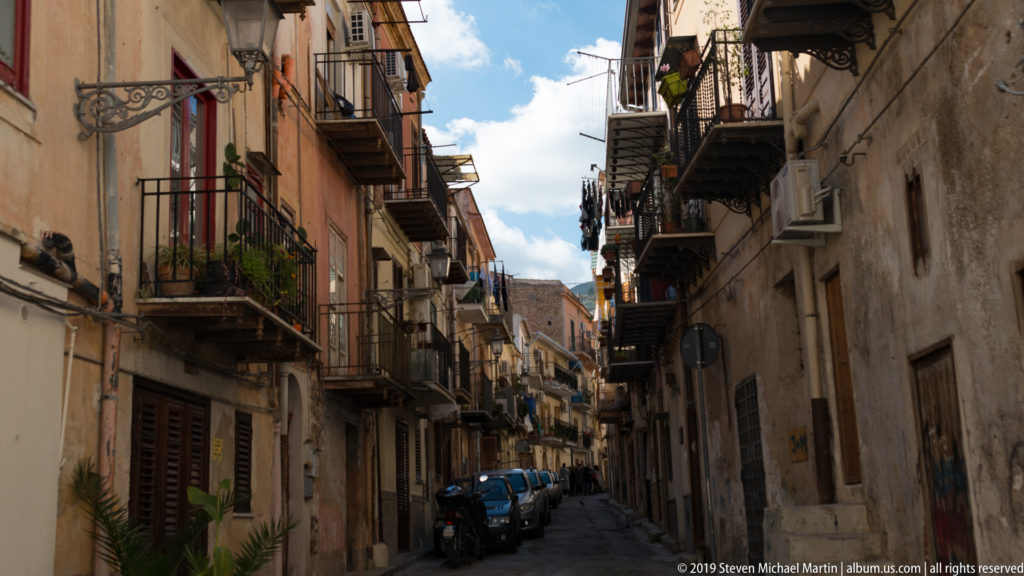 A narrow Monreale street lined with close-set buildings, balconies, and a parked blue car.