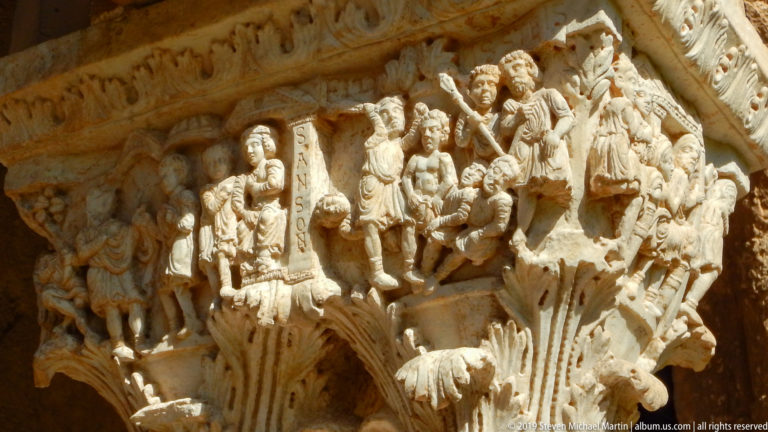 A stone capital with carved figures and a narrow vertical panel of lettering.