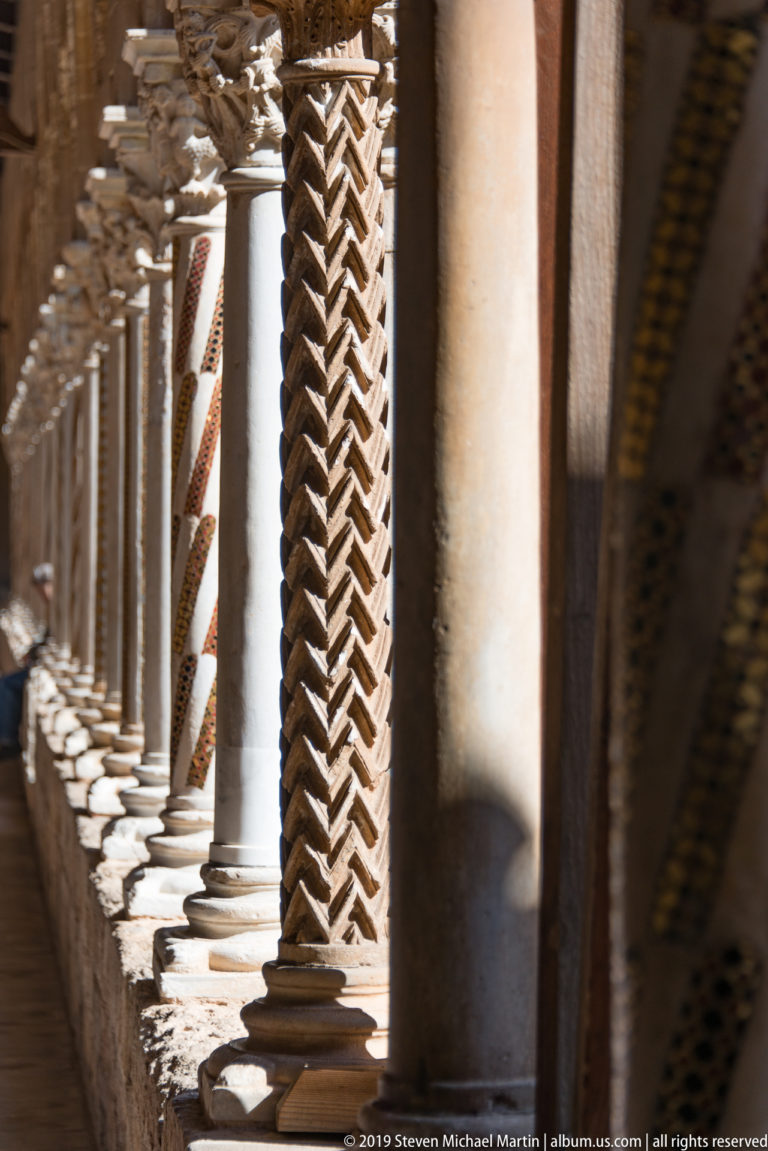 A deeply zigzag-carved column stands among plain shafts and repeating cloister arches.