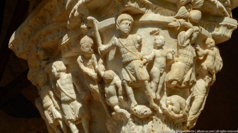 Carved robed figures fill a stone capital, with one figure elevated above a group.