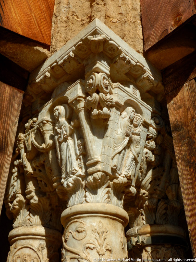 An upward view of a carved capital with small figures and rounded forms beneath its projecting edge.
