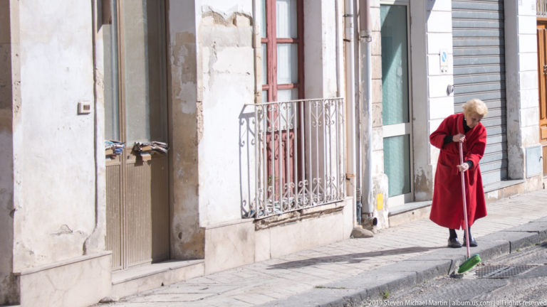 Older woman in a red coat sweeps a stone sidewalk with a green broom beside weathered residential façades.