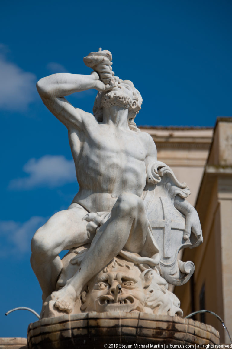 White stone fountain sculpture of a winged male figure riding a large sea creature against a deep blue sky.