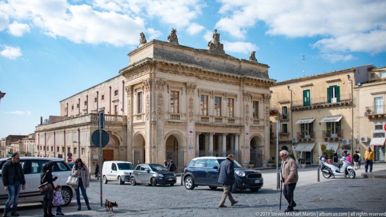 Ornate historic palazzo with three ground-floor arches, carved upper façade, rooftop statues, cars, scooters, and pedestrians.
