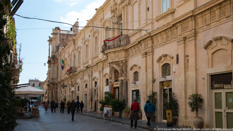 Pedestrians on a narrow Noto street lined with Baroque façades, balconies, shops, planters, and café seating.