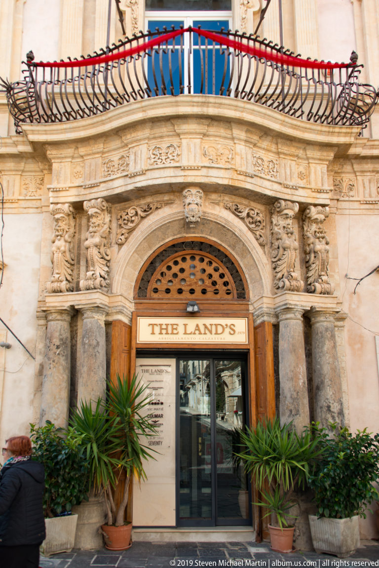 Ornate carved limestone doorway labeled “THE LAND’S” beneath a curved iron balcony, with potted plants at the entrance.
