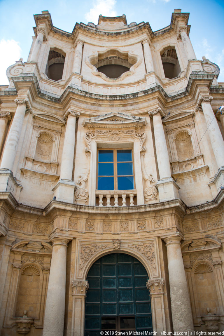 Low-angle view of an ornate curving Baroque church façade with columns, carved reliefs, central window, and dark doorway.