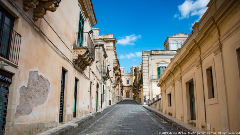Narrow uphill stone street between pale historic buildings with balconies, weathered plaster, and blue sky overhead.
