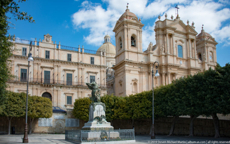 Noto Cathedral behind clipped trees and a foreground monument, with an adjoining palazzo and street lamps.