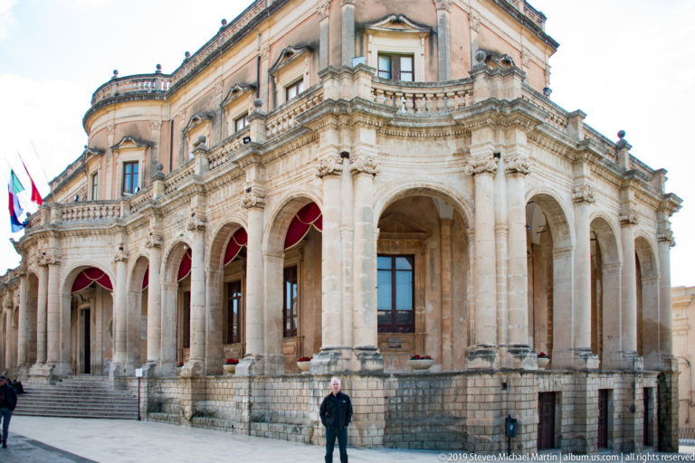 Oblique close view of Palazzo Ducezio’s arcaded corner with columns, red awnings, balustrades, and a pedestrian.