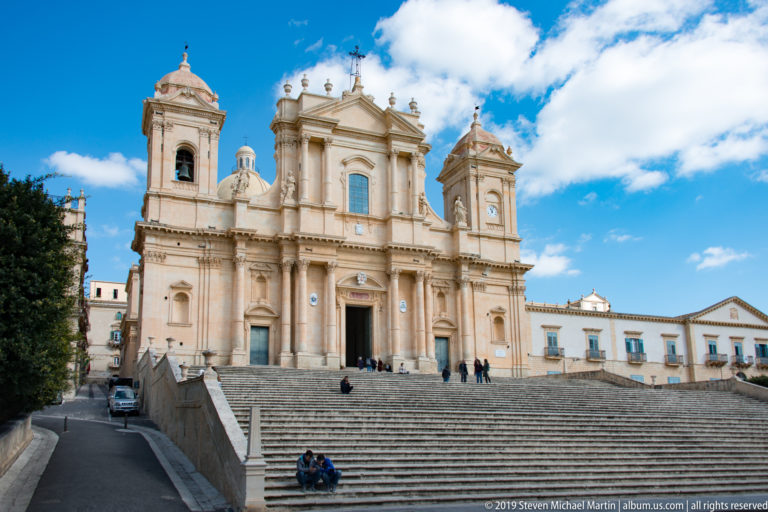 Noto Cathedral seen from the left approach, with broad stairs, retaining wall, twin towers, and people on the steps.
