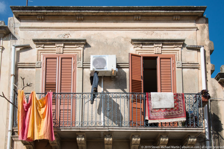 Colorful laundry and a patterned textile hang from a wrought-iron balcony between shuttered windows on a weathered façade.