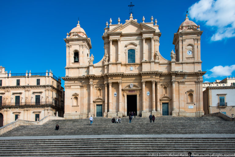 Wide frontal view of Noto Cathedral above broad steps, with twin towers, clocks, statues, and scattered visitors.