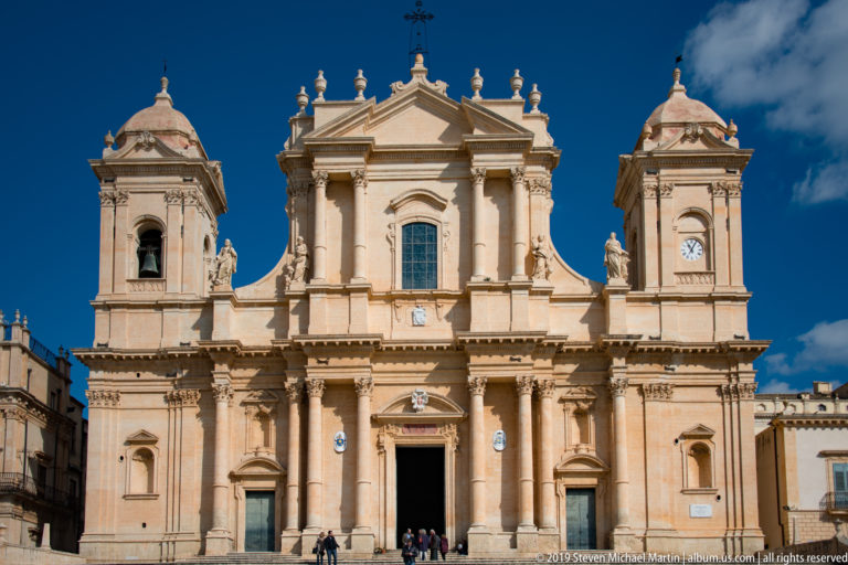 Tight frontal view of Noto Cathedral’s twin-towered Baroque façade with clocks, statues, columns, and open central door.