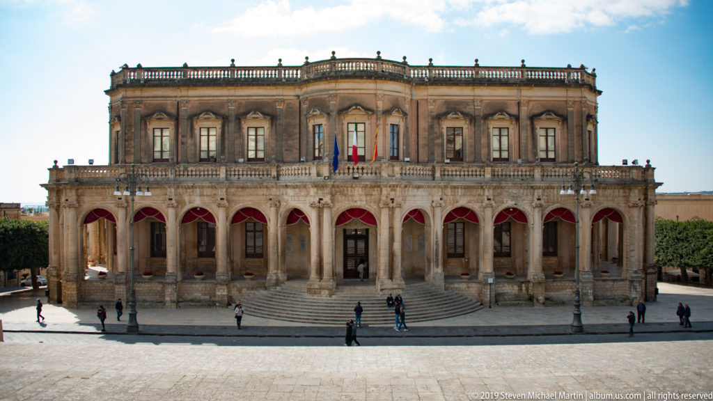 Frontal view of Palazzo Ducezio with long arcade, red awnings, flags, upper windows, and balustraded roofline.