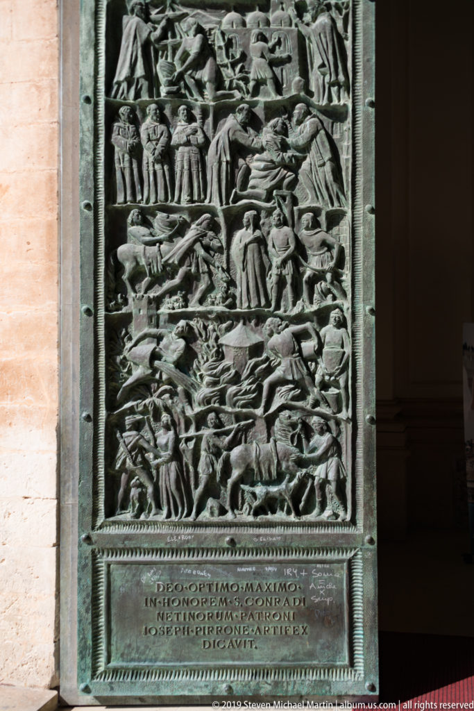 Full bronze door panel with crowded narrative relief above a rectangular Latin inscription on Noto Cathedral.