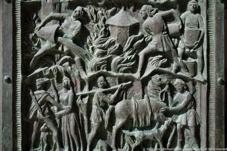 Close view of a bronze cathedral door relief crowded with human figures, animals, trees, and architectural details.