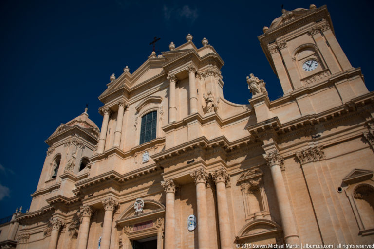 Oblique close view of Noto Cathedral’s upper façade, columns, statues, pediment, and right clock tower.