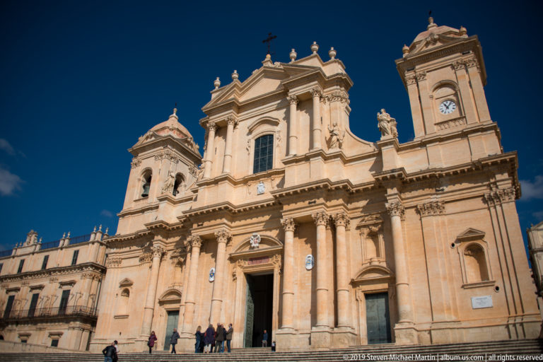 Low-angle frontal view of Noto Cathedral with twin bell towers, clocks, columns, statues, and visitors at the entrance.
