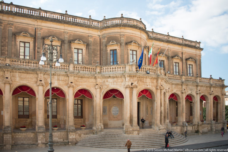 Palazzo Ducezio with arcaded loggia, red awnings, flags, balustrades, and broad steps facing the square.